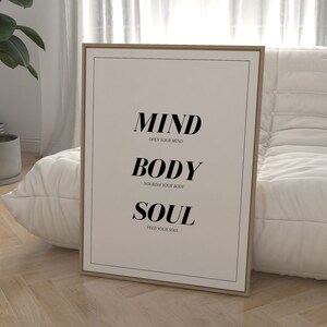 May include: A framed print with the words "MIND, BODY, SOUL" in black text on a white background. The text is centered on the print and each word is on a separate line. Below each word is a smaller line of text that says "OPEN YOUR MIND", "NOURISH YOUR BODY", and "FEED YOUR SOUL".