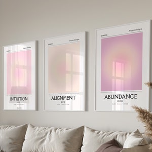May include: Three framed art prints with gradient backgrounds in pink, peach, and purple hues. The prints are labeled "Intuition," "Alignment," and "Abundance" with corresponding numbers. The artwork hangs above a beige sofa with decorative pillows.