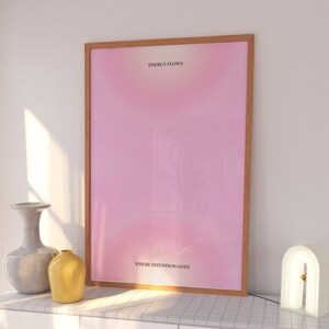 Puede incluir: Un póster rosa enmarcado con el texto "ENERGY FLOWS WHERE INTENTION GOES" colgado en una pared blanca sobre una cómoda blanca con dos cajones. Una lámpara blanca con un acento dorado está sobre la cómoda.