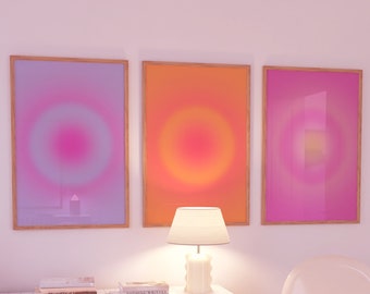 May include: Three framed abstract art prints with a gradient of pink, orange, and purple colors. The prints are hanging on a white wall above a white dresser with three drawers. A white lamp sits on the dresser. A white chair is in the foreground.