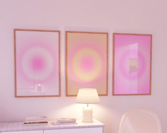 May include: Three framed abstract art prints with a gradient of pink, yellow, and white colors. The prints are hanging on a white wall above a white dresser with three drawers. A white lamp sits on the dresser. A white chair is in the foreground.