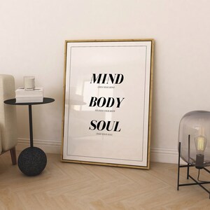 May include: A framed print with the words "MIND, BODY, SOUL" in black text on a white background. The text is centered on the print and each word is on a separate line. Below each word is a smaller line of text that reads "OPEN YOUR MIND", "NOURISH YOUR BODY", and "FEED YOUR SOUL".