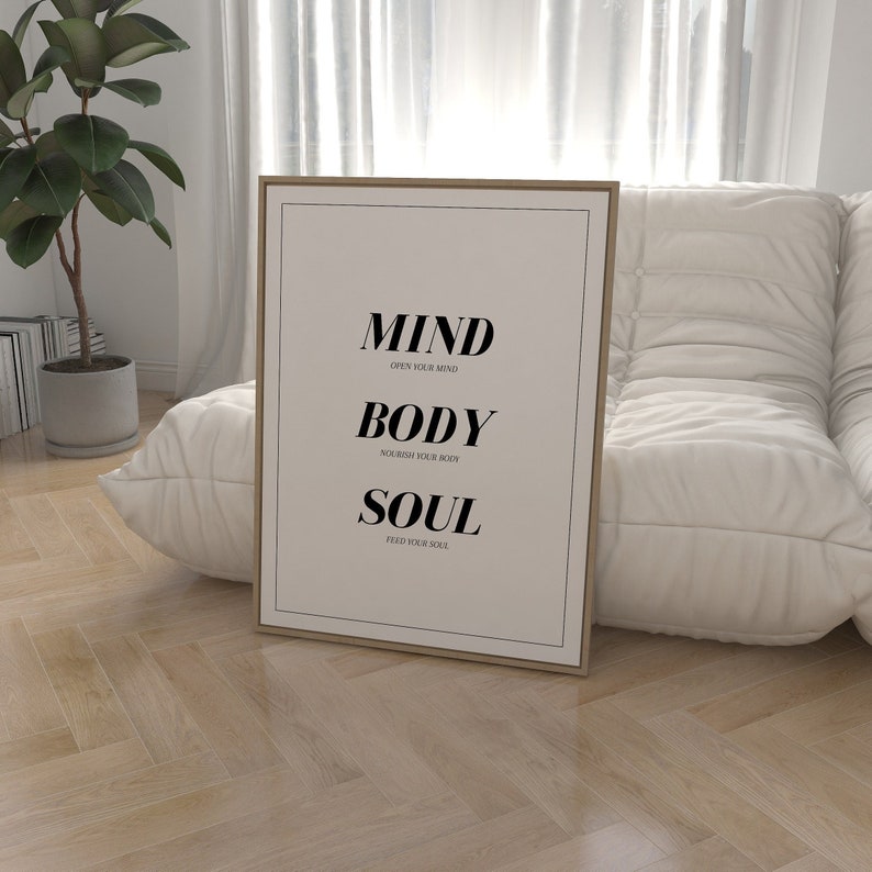 May include: A framed print with the words "MIND, BODY, SOUL" in black text on a white background. The text is centered on the print and each word is on a separate line. Below each word is a smaller line of text that says "OPEN YOUR MIND", "NOURISH YOUR BODY", and "FEED YOUR SOUL".