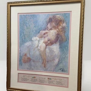 May include: Framed print featuring a mother cradling a sleeping baby, rendered in soft pastel colors. The artwork is set within a gold-toned frame and a pink mat. The print includes a poem at the bottom.