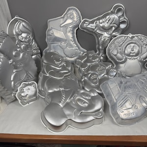 May include: A collection of vintage aluminum character cake pans, including Mickey Mouse, Big Bird, Winnie the Pooh, and a Cabbage Patch Kid. The pans are silver and in various shapes and sizes, perfect for baking themed cakes.