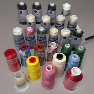 May include: A collection of spools of thread in various colors, including black, white, pink, green, and yellow. Some spools are labeled "SureLock" and "Maxi Lock". The spools are cylindrical and have a white or black top.