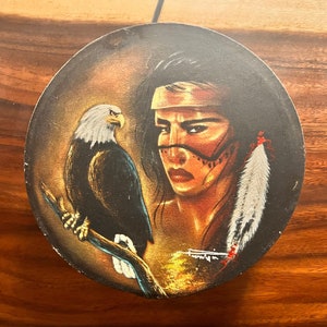 May include: A circular painting depicting a Native American woman with a white feather headdress and a bald eagle perched on a branch. The woman has dark hair and brown eyes, and she is wearing a beaded necklace. The background is a dark brown colour.