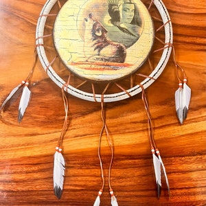 Peut inclure: Un attrape-rêves blanc avec des cordons en cuir brun et des plumes. Le centre de l'attrape-rêves présente une image imprimée d'un loup hurlant à la lune et d'une femme amérindienne avec une coiffe.