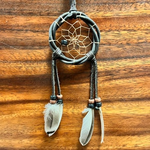 May include: A small, handcrafted dreamcatcher with a dark gray woven hoop and a web of tan string. Two gray and white feathers dangle below, accented with wooden beads. The dreamcatcher is suspended by a dark gray cord, with a tag that says "MEXICO".