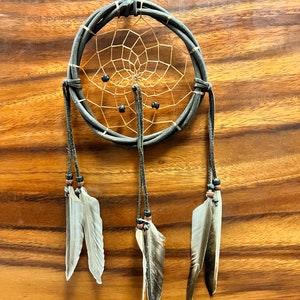 May include: A handcrafted dreamcatcher with a dark gray woven frame and a tan web. The dreamcatcher features three hanging strands with feathers and beads. The background is a wooden surface.