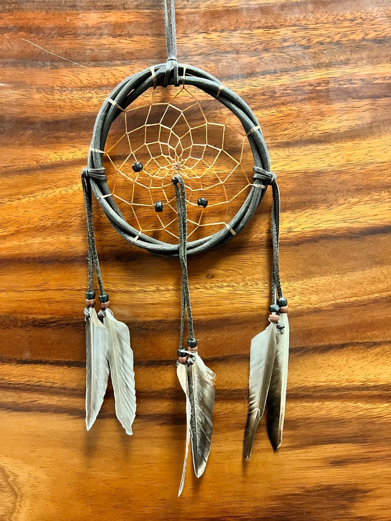 May include: A handcrafted dreamcatcher with a dark gray woven frame and a tan web. The dreamcatcher features three hanging strands with feathers and beads. The background is a wooden surface.