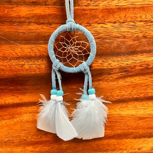 May include: A light blue dreamcatcher with white feathers and blue beads hanging from it. The dreamcatcher is made of suede and has a brown leather cord attached to it. The tag on the cord reads "Mexico".