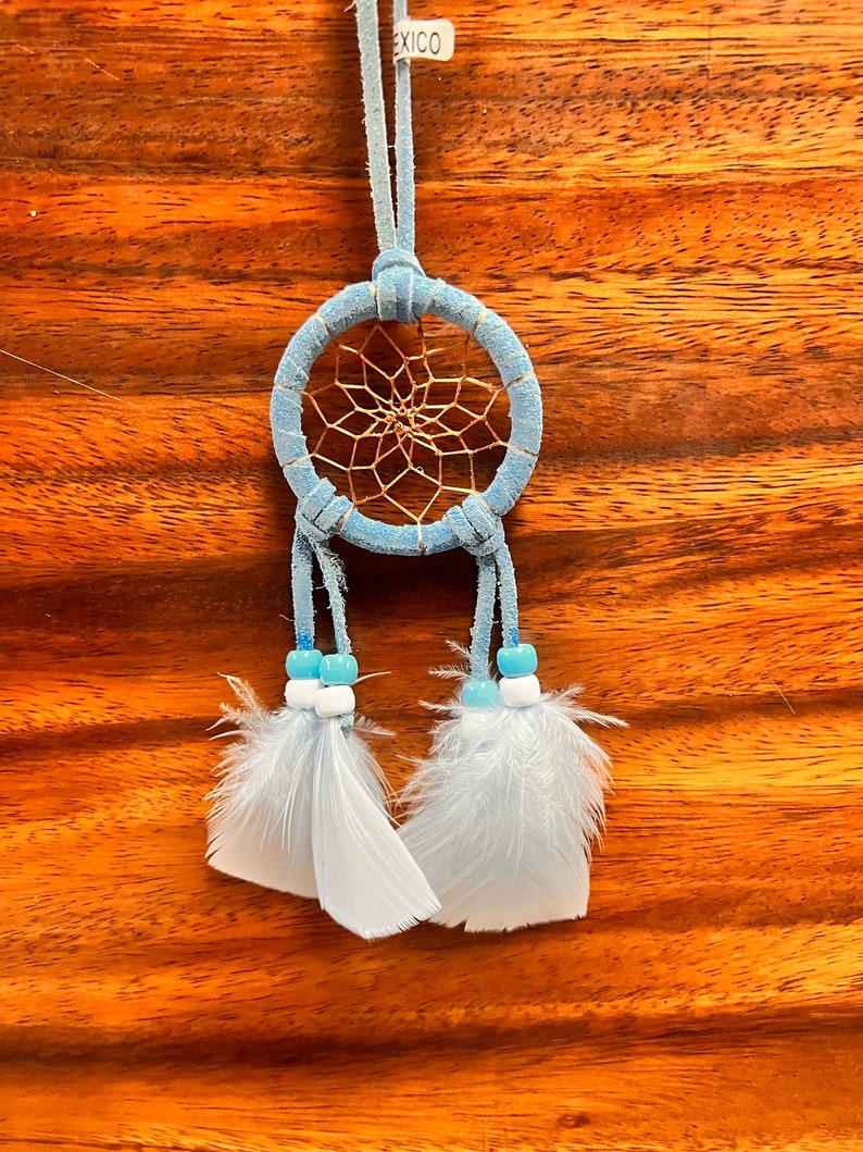 May include: A light blue dreamcatcher with white feathers and blue beads hanging from it. The dreamcatcher is made of suede and has a brown leather cord attached to it. The tag on the cord reads "Mexico".