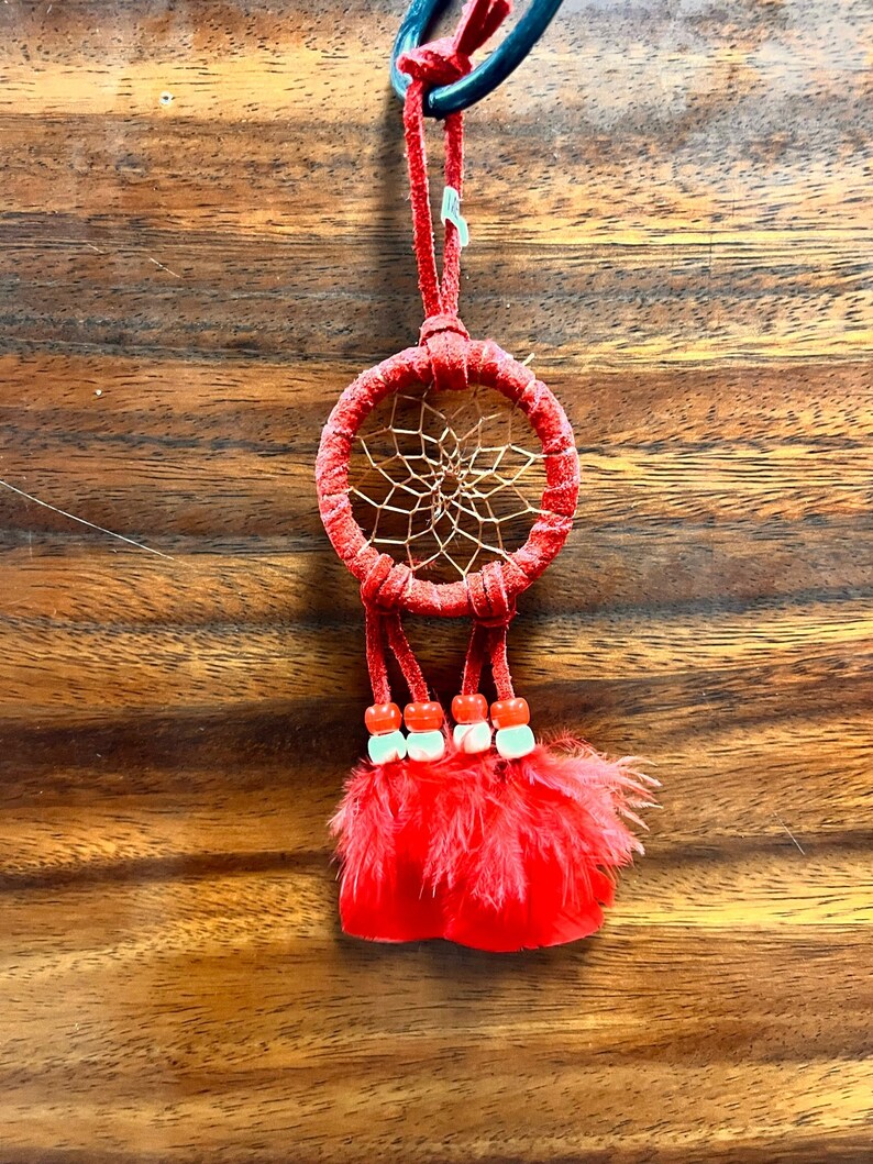 May include: A red dreamcatcher with a beaded tassel and red feathers. The dreamcatcher is hanging from a black cord.