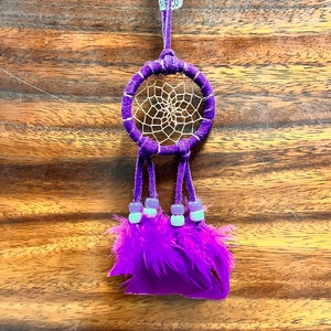 May include: Purple dream catcher with white beads and feathers hanging from a purple cord. The dream catcher has a white web design and the tag "MEXCO" is attached to the cord.