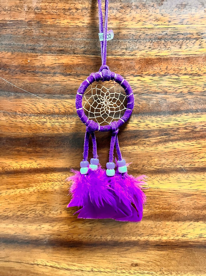 May include: Purple dream catcher with white beads and feathers hanging from a purple cord. The dream catcher has a white web design and the tag "MEXCO" is attached to the cord.