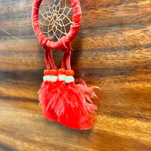 May include: A red dreamcatcher with a brown web and red feathers hanging from it. The dreamcatcher is hanging from a wooden surface.