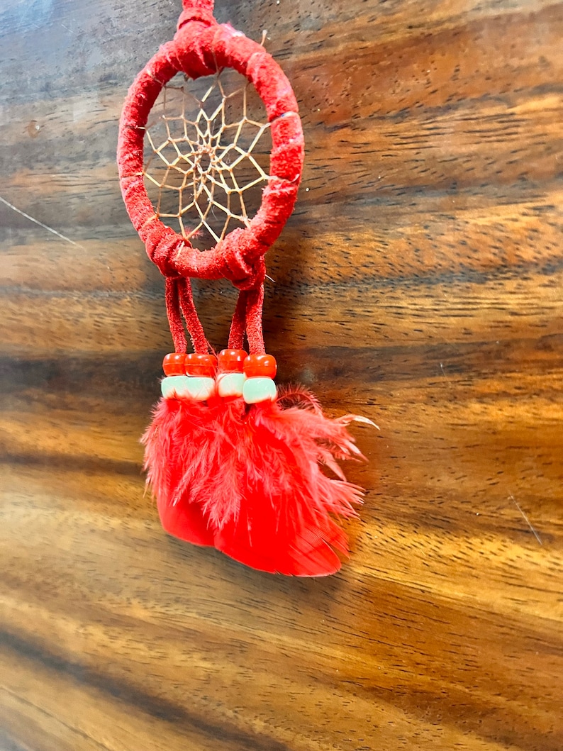 May include: A red dreamcatcher with a brown web and red feathers hanging from it. The dreamcatcher is hanging from a wooden surface.