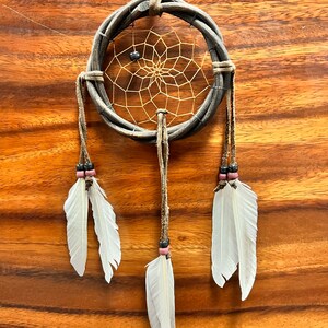 May include: A handcrafted dreamcatcher with a woven gray hoop and a delicate web center. White feathers hang from the bottom, accented with beads. The dreamcatcher is suspended by a leather cord, perfect for wall decor.