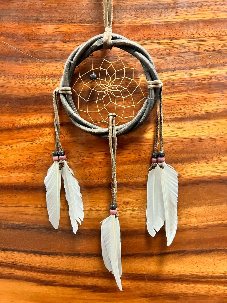 May include: A handcrafted dreamcatcher with a woven gray hoop and a delicate web center. White feathers hang from the bottom, accented with beads. The dreamcatcher is suspended by a leather cord, perfect for wall decor.