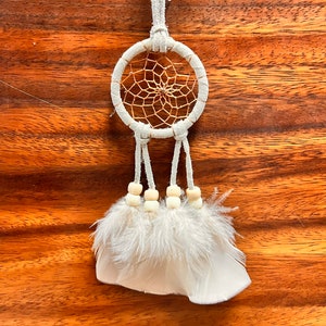 May include: White dream catcher with a brown woven web and white feathers hanging from it. The dream catcher is hanging from a white cord and has white wooden beads.