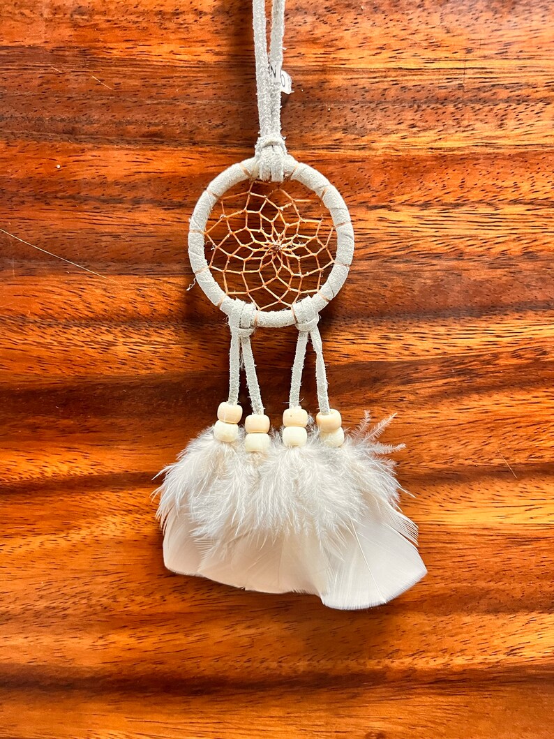 May include: White dream catcher with a brown woven web and white feathers hanging from it. The dream catcher is hanging from a white cord and has white wooden beads.