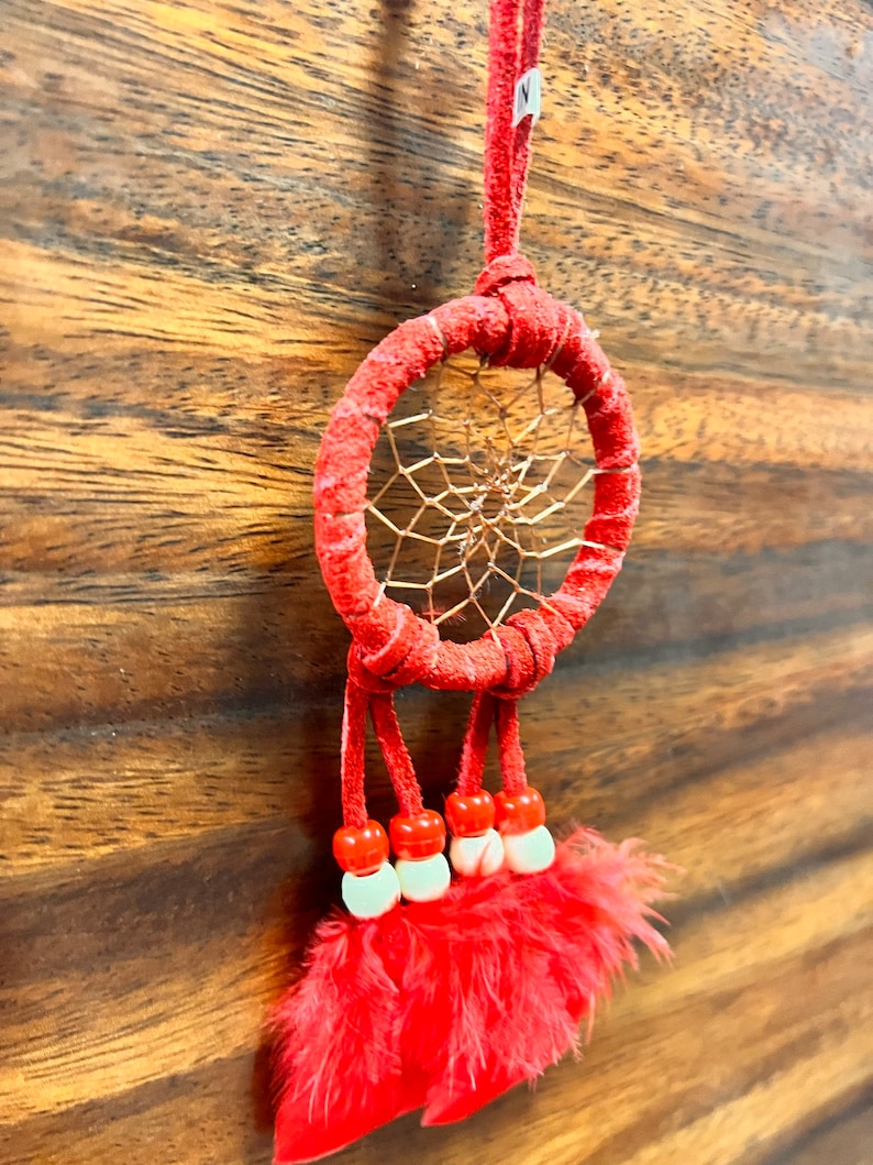 May include: Red dreamcatcher with a copper wire web and red feathers. The dreamcatcher has a red suede cord and white beads.