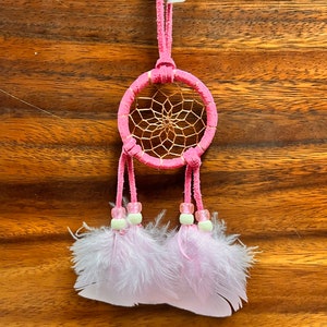 May include: Pink dream catcher with white feathers and beads. The dream catcher has a pink suede cord and a small tag that says "XICO".