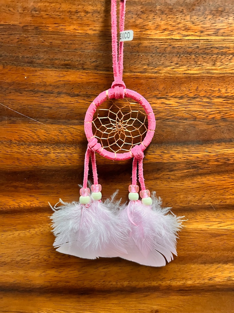 May include: Pink dream catcher with white feathers and beads. The dream catcher has a pink suede cord and a small tag that says "XICO".