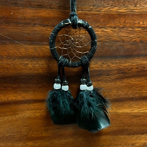 May include: A black dreamcatcher with black feathers and white beads hanging from it. The dreamcatcher is hanging from a black cord.