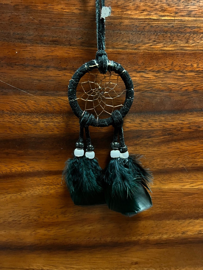 May include: A black dreamcatcher with black feathers and white beads hanging from it. The dreamcatcher is hanging from a black cord.