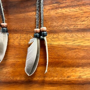 May include: Close-up of a handcrafted dreamcatcher detail. Features gray and white feathers, wooden beads, and braided leather cords. The background is a warm-toned wooden surface. The dreamcatcher is designed with natural elements.