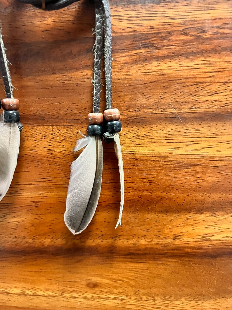 May include: Close-up of a handcrafted dreamcatcher detail. Features gray and white feathers, wooden beads, and braided leather cords. The background is a warm-toned wooden surface. The dreamcatcher is designed with natural elements.