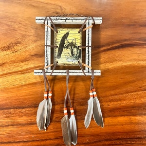 K&ouml;nnte beinhalten: Ein kleiner Holzrahmen mit einer wei&szlig;en Lackierung, der einen Druck eines Wolfes und eines Vogels zeigt. Der Rahmen ist mit braunen Lederriemen und Federn verziert, was ein indianisch inspiriertes Design ergibt.