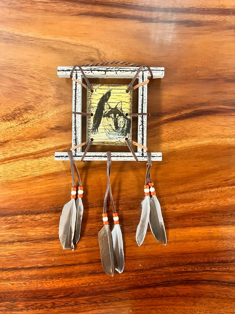 K&ouml;nnte beinhalten: Ein kleiner Holzrahmen mit einer wei&szlig;en Lackierung, der einen Druck eines Wolfes und eines Vogels zeigt. Der Rahmen ist mit braunen Lederriemen und Federn verziert, was ein indianisch inspiriertes Design ergibt.