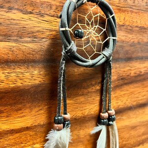 May include: A handcrafted dreamcatcher with a woven black frame and a delicate web design. The dreamcatcher features a black bead in the center and is adorned with hanging leather strands, wooden beads, and soft white feathers. The background is a wood grain.