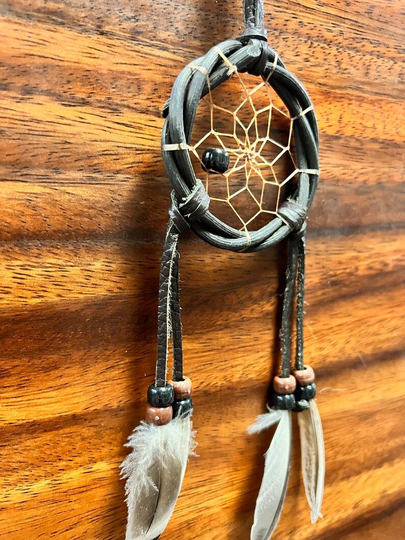 May include: A handcrafted dreamcatcher with a woven black frame and a delicate web design. The dreamcatcher features a black bead in the center and is adorned with hanging leather strands, wooden beads, and soft white feathers. The background is a wood grain.