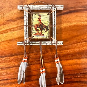K&ouml;nnte beinhalten: Ein wei&szlig;er Holzrahmen mit einem Bild eines amerikanischen Ureinwohners, der ein Pferd reitet. Der Rahmen ist mit braunen Lederriemen und drei wei&szlig;en Federn mit orangefarbenen Perlen verziert.