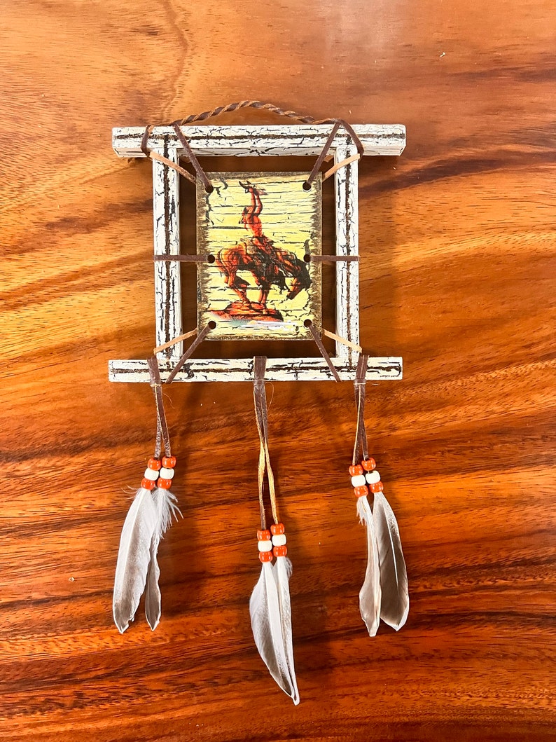 K&ouml;nnte beinhalten: Ein wei&szlig;er Holzrahmen mit einem Bild eines amerikanischen Ureinwohners, der ein Pferd reitet. Der Rahmen ist mit braunen Lederriemen und drei wei&szlig;en Federn mit orangefarbenen Perlen verziert.
