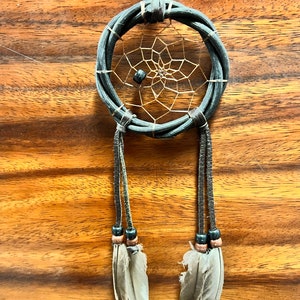 May include: A handcrafted dreamcatcher with a woven dark gray ring, a tan web, and hanging leather strands. The strands are adorned with beads and feathers. The dreamcatcher is suspended against a wooden background.