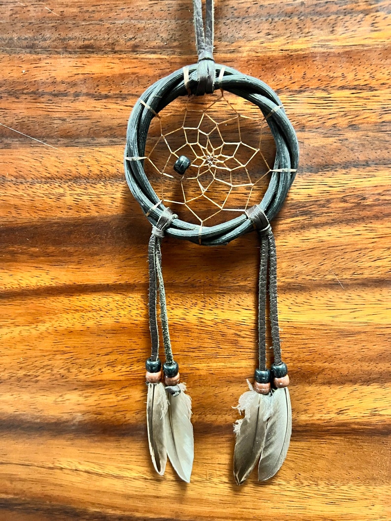 May include: A handcrafted dreamcatcher with a woven dark gray ring, a tan web, and hanging leather strands. The strands are adorned with beads and feathers. The dreamcatcher is suspended against a wooden background.