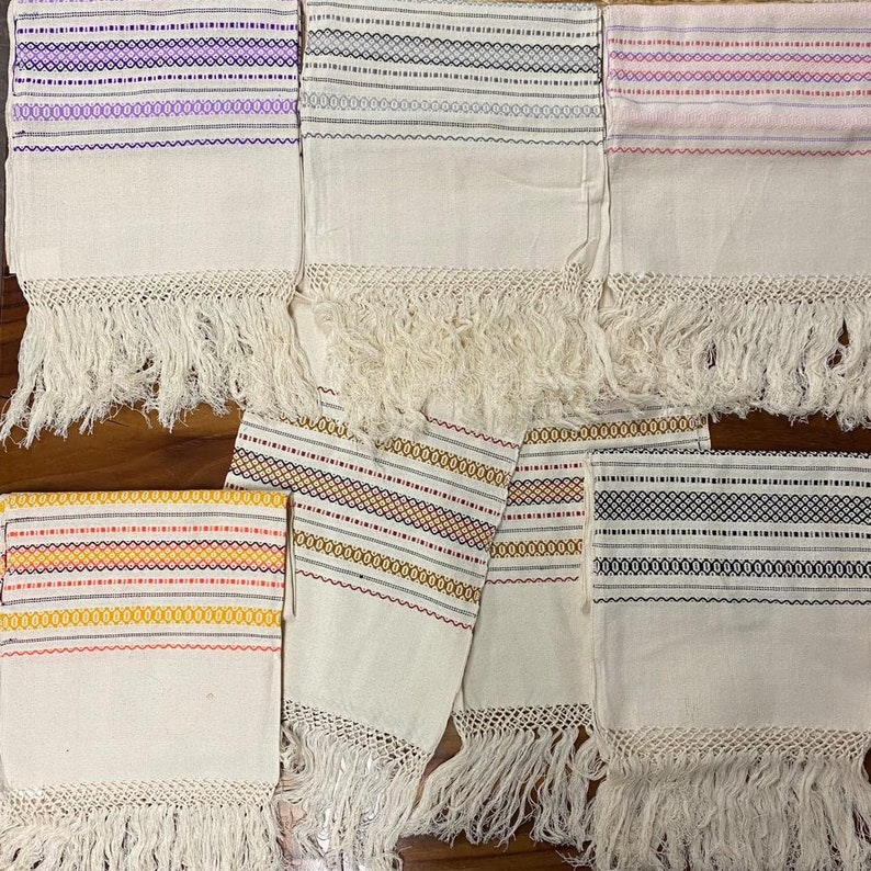May include: Six white cotton tablecloths with colourful woven stripes and fringe. The tablecloths are arranged in two rows of three. The tablecloths have a variety of colours including purple, grey, pink, yellow, red, and black.