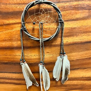 May include: A handcrafted dreamcatcher with a woven gray hoop and a delicate web design. Three strands hang down, each adorned with feathers and beads. The dreamcatcher is suspended against a wooden background.