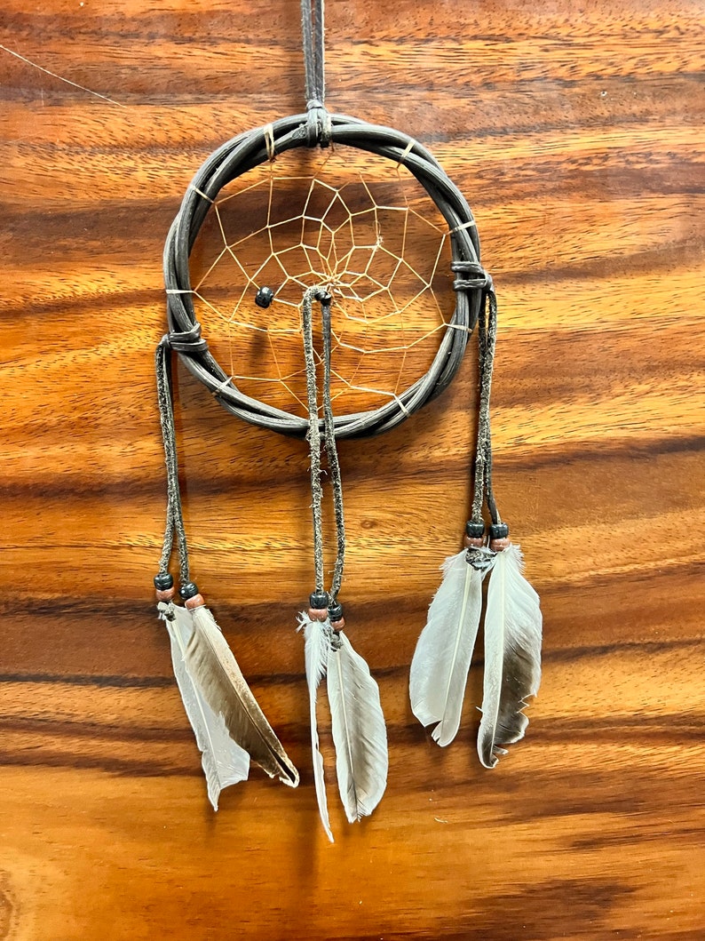 May include: A handcrafted dreamcatcher with a woven gray hoop and a delicate web design. Three strands hang down, each adorned with feathers and beads. The dreamcatcher is suspended against a wooden background.