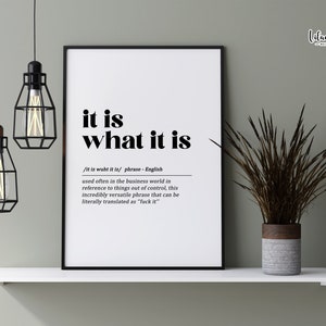 May include: A black and white printable wall art print with the phrase "it is what it is" and a definition of the phrase. The print is framed and hanging on a white shelf with two black caged light bulbs hanging above it.