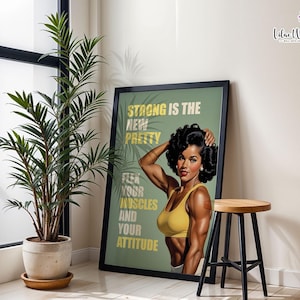 May include: A framed wall art print featuring a muscular person with dark hair wearing a yellow sports bra. The text reads: STRONG IS THE NEW PRETTY, FLEX YOUR MUSCLES AND YOUR ATTITUDE. The background is sage green with palm leaf silhouettes.