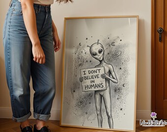 May include: A framed wall art print featuring a black and white illustration of an alien holding a sign that reads I DON'T BELIEVE IN HUMANS. The artwork includes cosmic details like stars and spiral galaxies set against a stippled background.