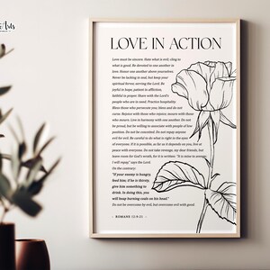 Puede incluir: Impresión enmarcada con la frase "LOVE IN ACTION" y una ilustración floral. El texto incluye un pasaje de Romanos 12:9-21. La obra de arte es en blanco y negro, con un marco de madera clara.