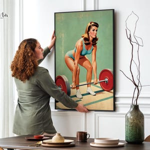 May include: A vintage style poster featuring a woman lifting a barbell. The woman is wearing a blue and white swimsuit and has blonde hair. The background is a light green color. The poster is framed in black.