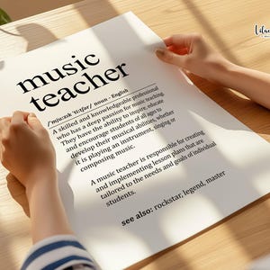 May include: A white print with the words "music teacher" in a bold, black font. The definition of a music teacher is printed below the title. The print is held by a person on a wooden surface.